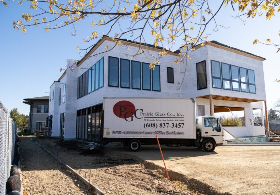 A two-story modern house under construction with large windows, beside a Prairie Glass Co. truck parked on a dirt driveway under a tree with autumn leaves.
