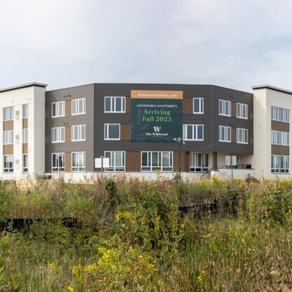 A modern three-story apartment complex with a sign announcing "New Apartments Arriving Fall 2023," surrounded by overgrown grass and wild plants under a cloudy sky.