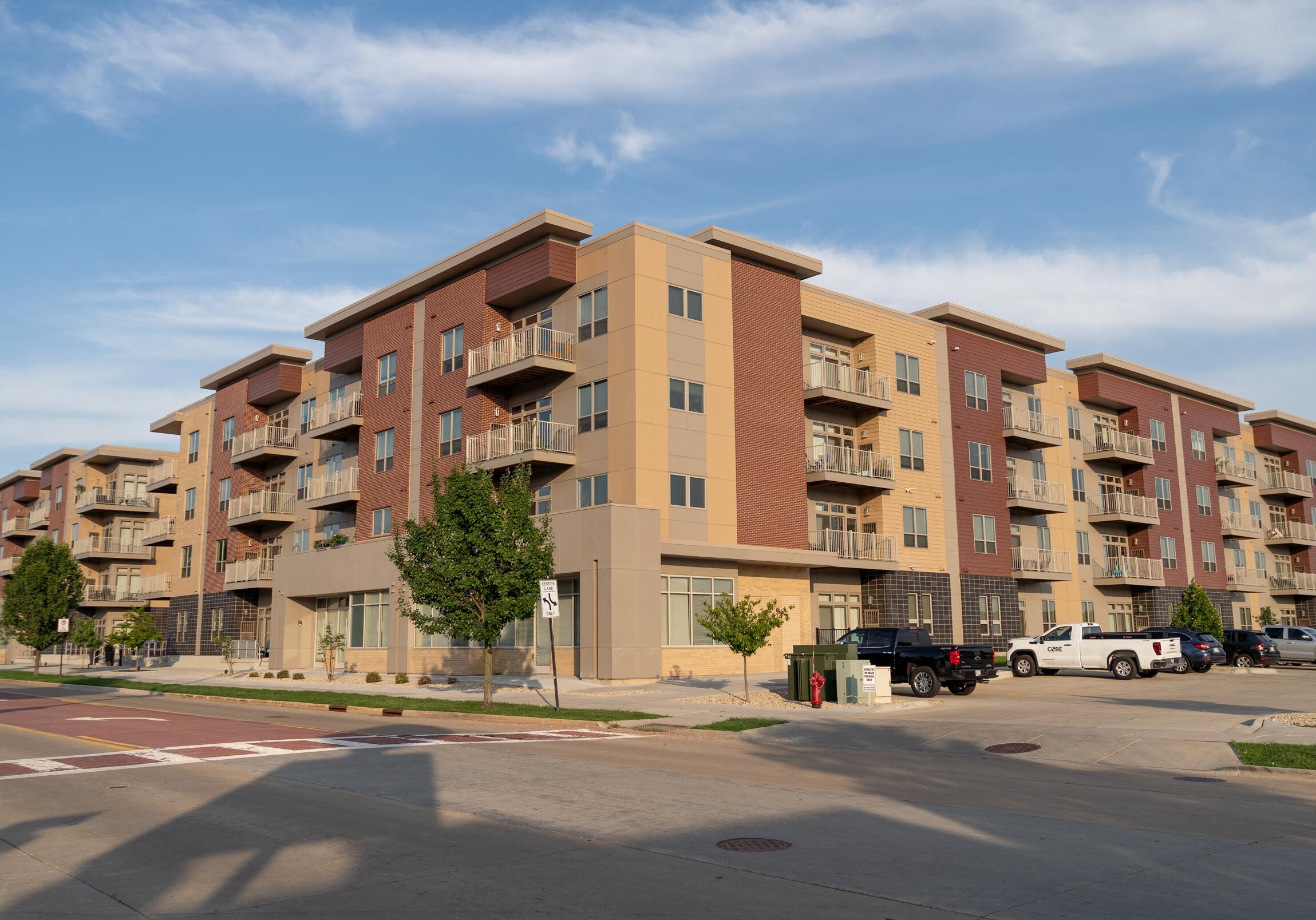 A modern four-story apartment building with balconies and parked cars in front, set on a sunny day with a partly cloudy sky.