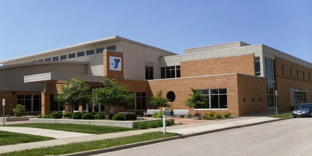 A modern brick and glass building with a YMCA logo on the facade, surrounded by trees and a small parking area on a sunny day.