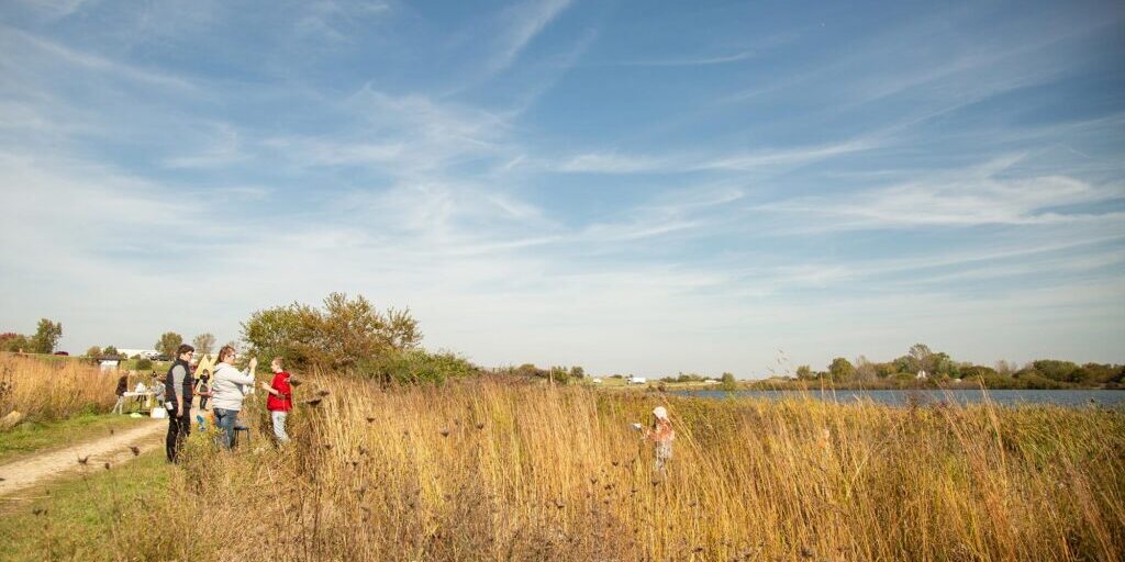 A group of people stand on a grassy path near tall dry grass and a body of water under a blue sky with wispy clouds.