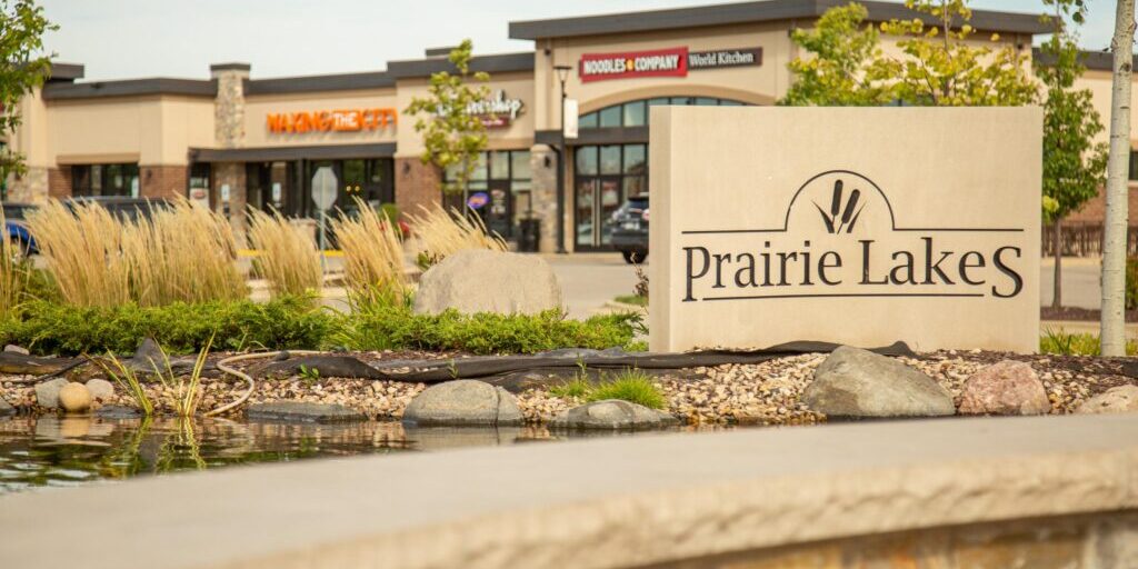 A stone sign reading "Prairie Lakes" stands by a small pond, with shops and restaurants visible in the background.