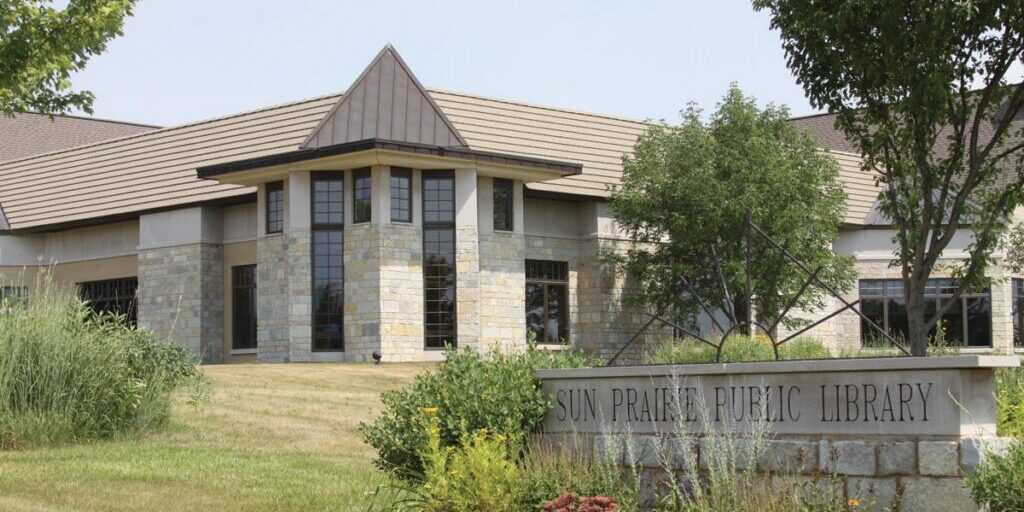 Exterior view of the Sun Prairie Public Library building, featuring stone walls, large windows, and landscaped greenery in front.