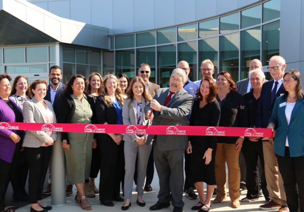 A group of people stand in front of a building for a ribbon-cutting ceremony. A man and woman hold large scissors, surrounded by other smiling participants.