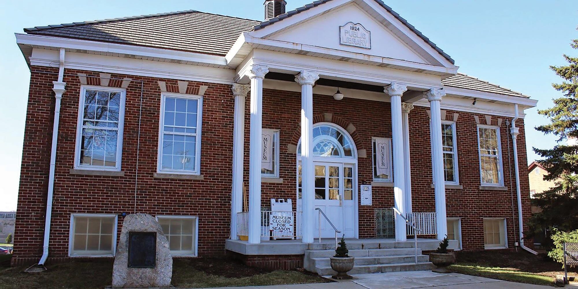 A red brick building with white columns and a triangular pediment, featuring large windows and a central arched doorway with steps leading to the entrance.