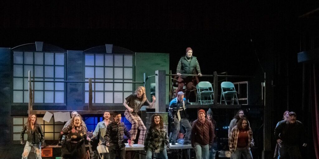 A group of performers stand and sing on a stage set resembling an urban loft, dressed in casual, winter clothing with a few props and chairs in the background.