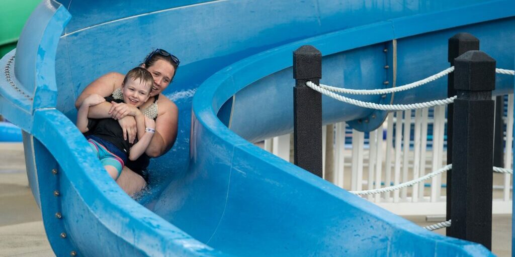 An adult and a child slide down a blue water slide together, with the child smiling and the adult holding onto them.