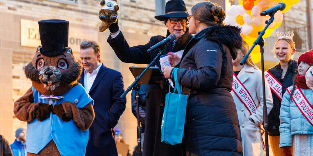 A man in a black hat holds up a stuffed animal on stage with a woman in a coat, a large groundhog mascot, and pageant titleholders at an outdoor event.