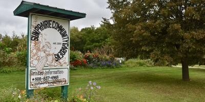 A sign for Sun Prairie Community Gardens stands on a grassy lawn, with trees and flowerbeds in the background under a cloudy sky.