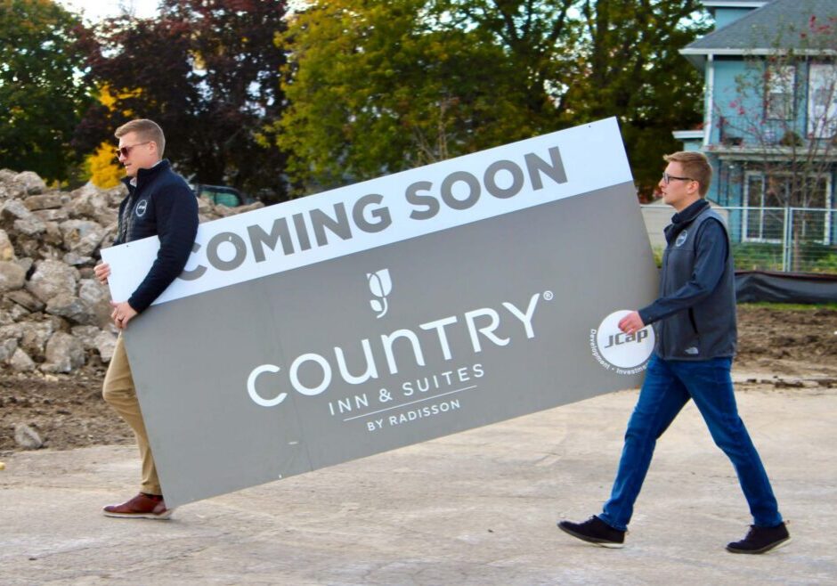 Two men carry a large "Coming Soon - Country Inn & Suites by Radisson" sign at a construction site with dirt and rocks in the background.