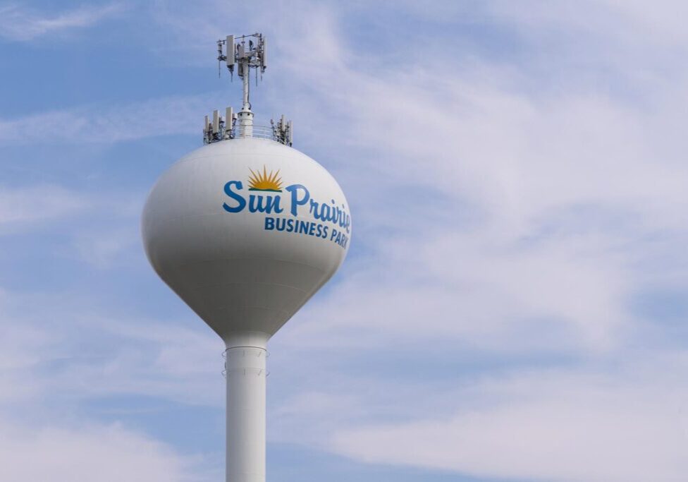 A white water tower with antennas and the text "Sun Prairie Business Park" stands against a blue sky with light clouds.